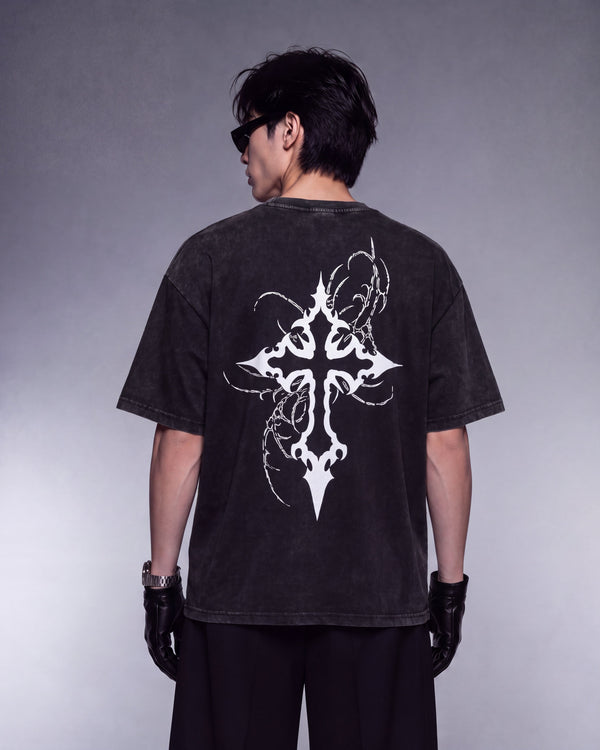 A person shown from the back wearing a faded black short-sleeve T-shirt with a large white ornate cross graphic on the back, paired with black pants and black gloves, against a gray studio background.