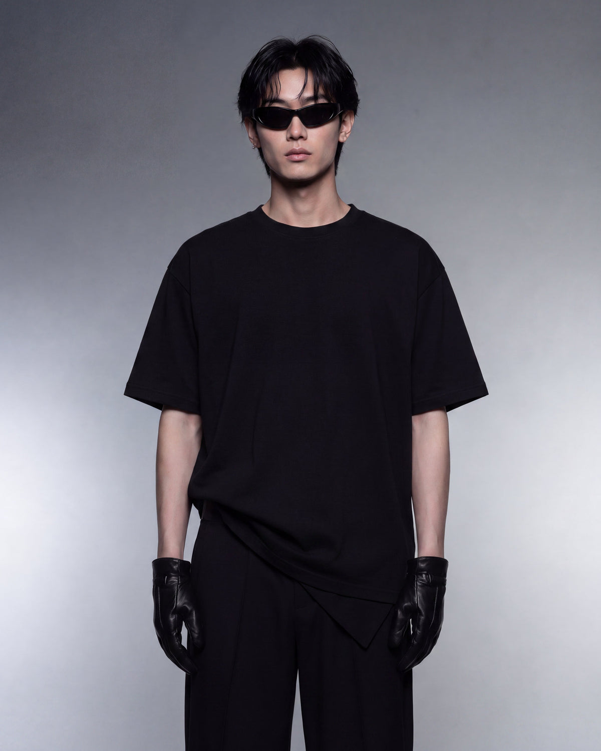 A model wearing a black oversized short-sleeve t-shirt, black pants, black leather gloves, and black sunglasses, standing against a gray studio background.