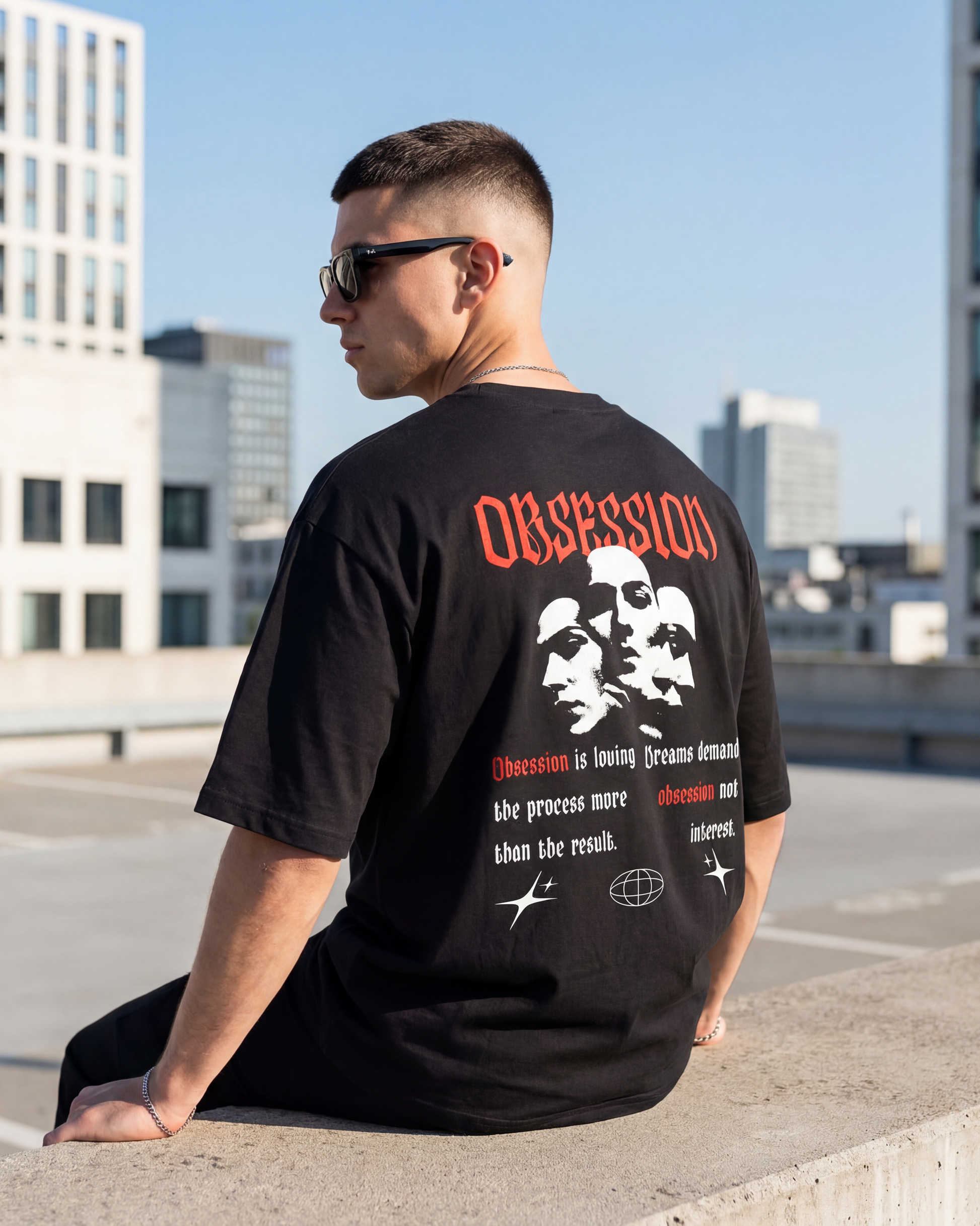 Man wearing a black t-shirt with graphic designs and text, sitting on a rooftop with buildings in the background.