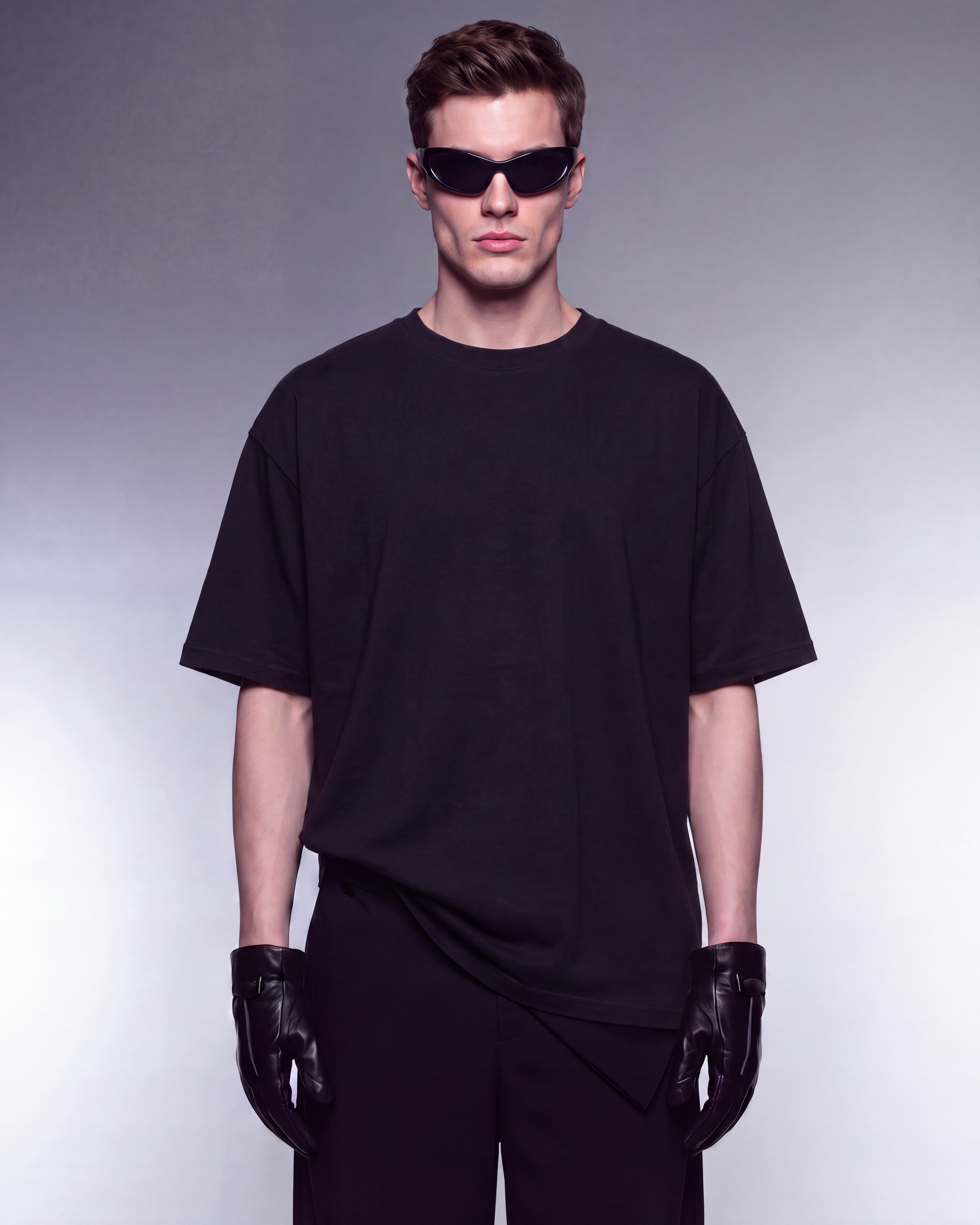 A man wearing an oversized black t-shirt, black pants, black gloves, and black sunglasses, standing against a gray gradient studio background.