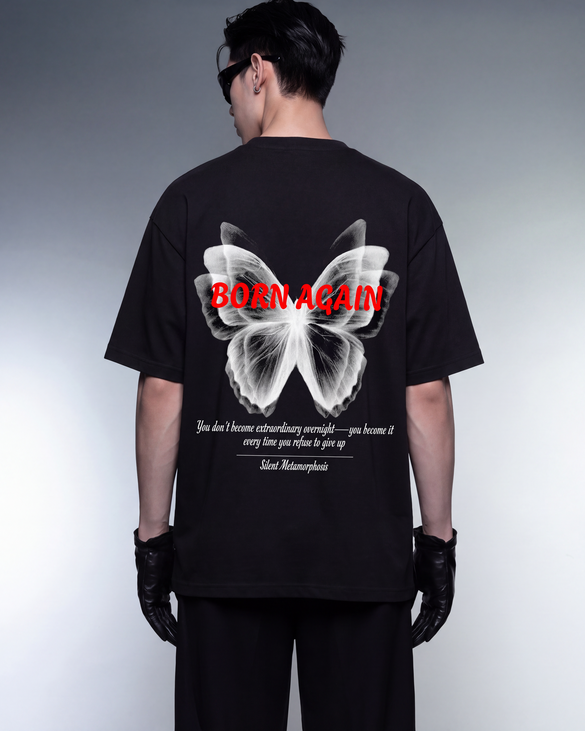 A model from behind wearing a black t-shirt with a large white butterfly graphic on the back, red 'BORN AGAIN' text, and a quote by Albert Einstein. The model is also wearing black pants and gloves against a gray studio background.