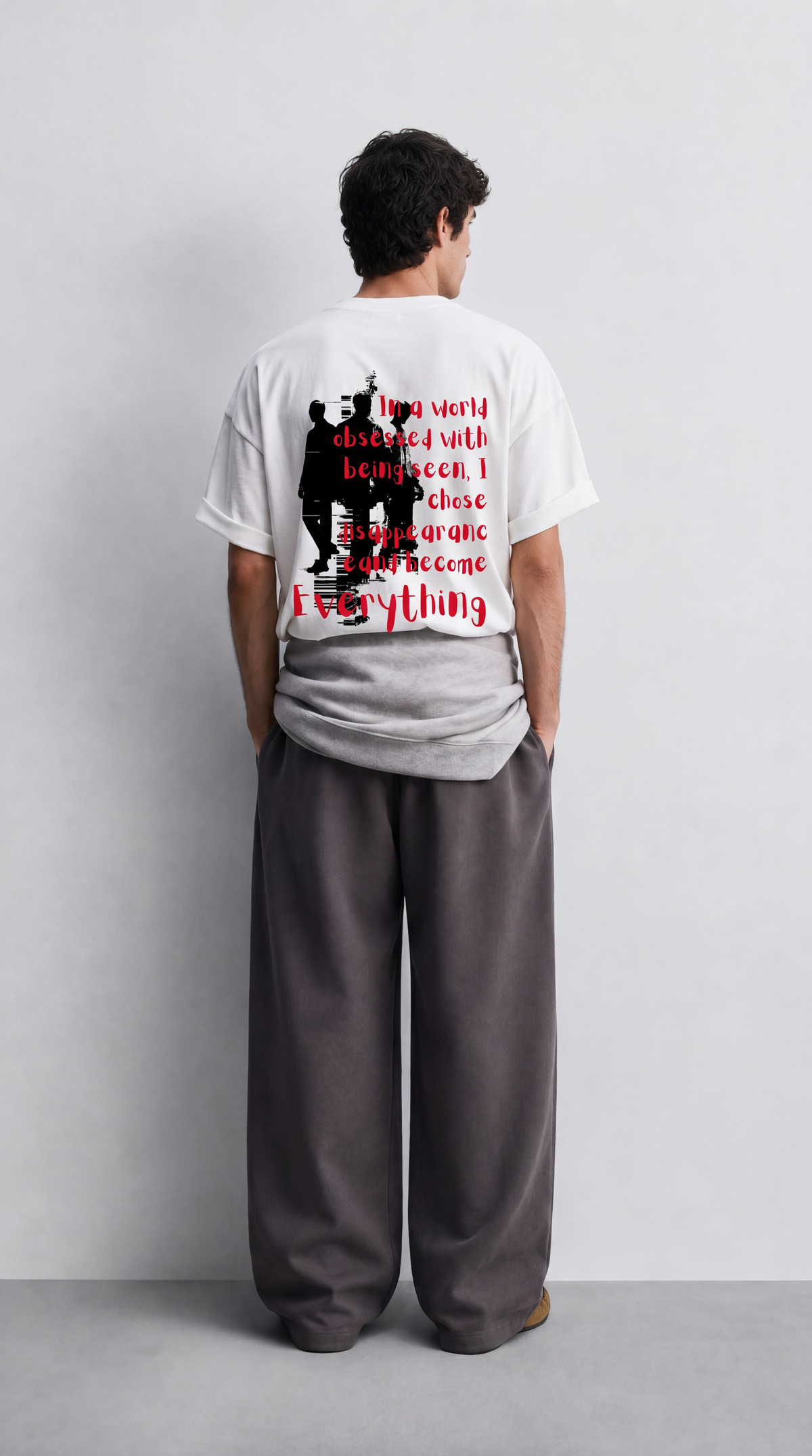 A man stands with his back to the camera wearing a white graphic t-shirt and loose dark gray sweatpants in a minimalist studio setting.