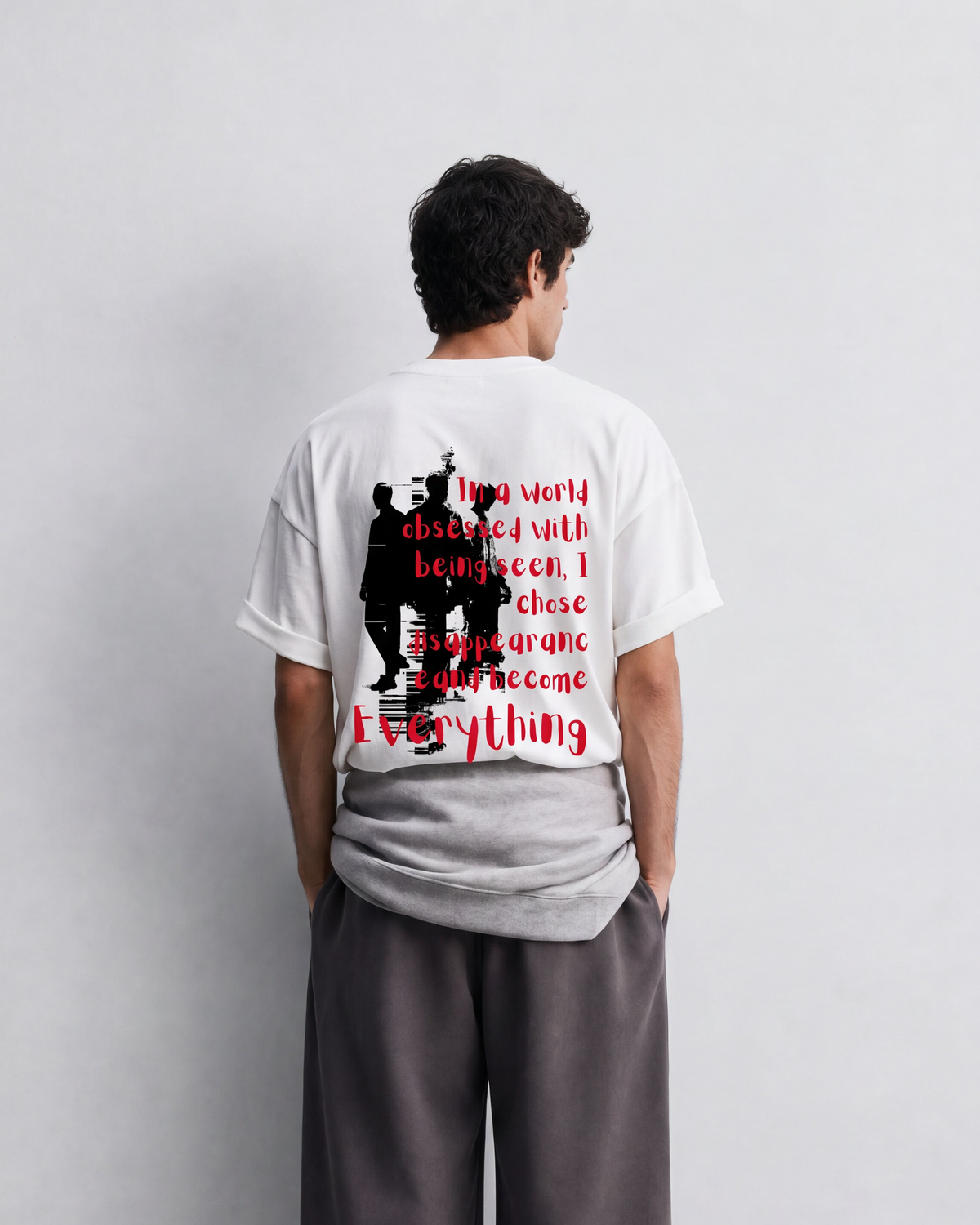 A man stands with his back to the camera wearing a white graphic t-shirt and loose dark gray sweatpants in a minimalist studio setting.