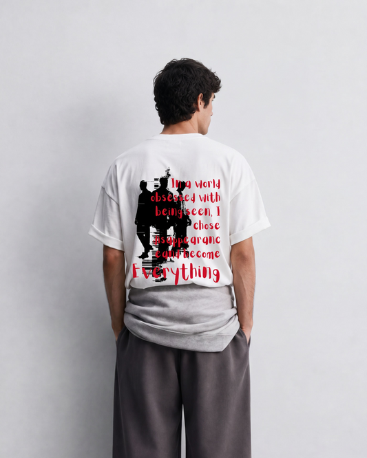 A man stands with his back to the camera wearing a white graphic t-shirt and loose dark gray sweatpants in a minimalist studio setting.