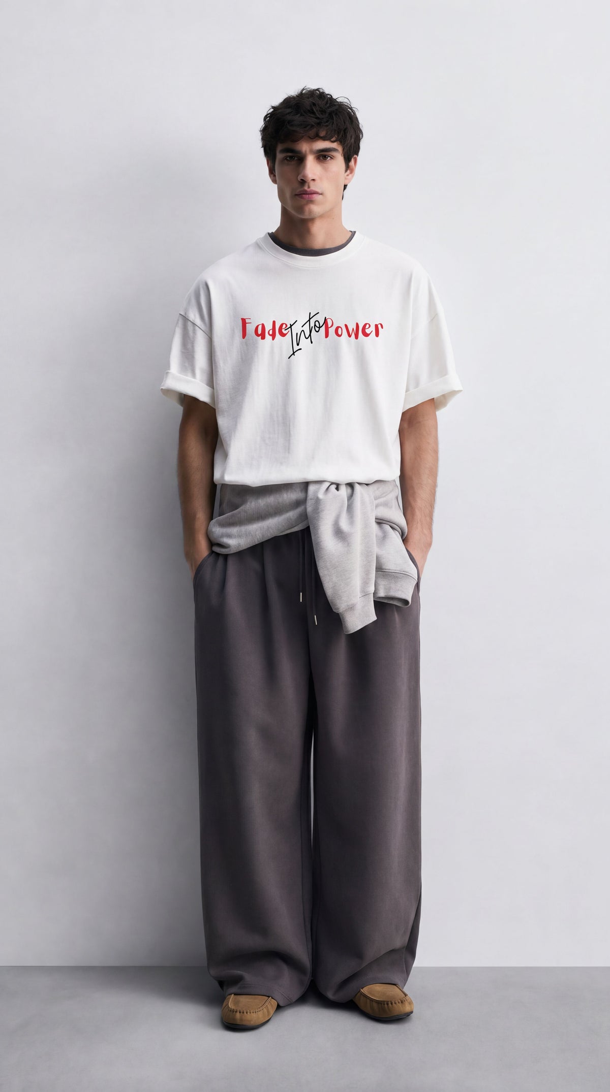 A man stands against a light gray wall wearing a white T-shirt with red and black text reading 'I love you ever', dark gray wide-leg pants, a light gray sweatshirt tied around his waist, and brown shoes.