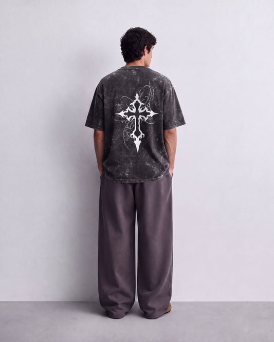 A man viewed from behind wearing a dark gray mineral-wash t-shirt with a large white tribal cross graphic on the back, paired with loose dark gray pants, standing against a light gray wall.