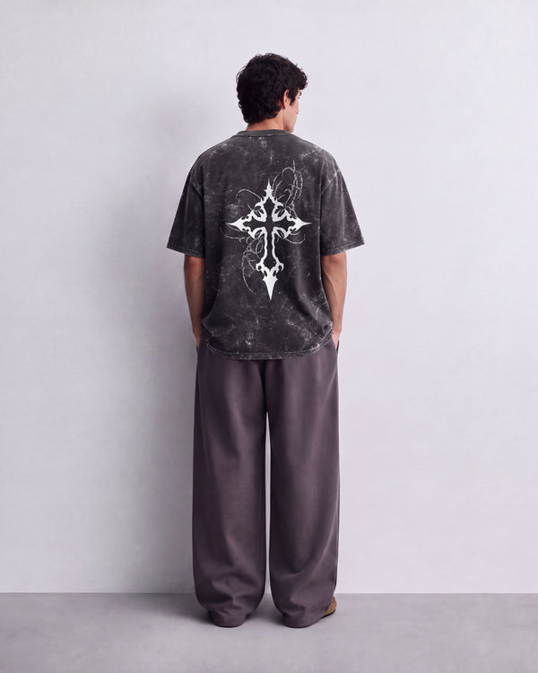 A man viewed from behind wearing a dark gray mineral-wash t-shirt with a large white tribal cross graphic on the back, paired with loose dark gray pants, standing against a light gray wall.