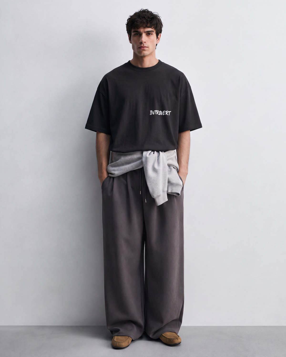 A man stands against a light gray studio wall wearing a black oversized T-shirt with a small white graphic on the chest, loose dark gray wide-leg pants, a light gray sweatshirt tied around his waist, and brown slip-on shoes.