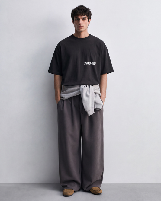 A man stands against a light gray studio wall wearing a black oversized T-shirt with a small white graphic on the chest, loose dark gray wide-leg pants, a light gray sweatshirt tied around his waist, and brown slip-on shoes.