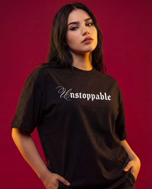 A woman modeling a black oversized t-shirt with the word 'Unstoppable' printed in a white gothic font across the chest, set against a solid dark red background.